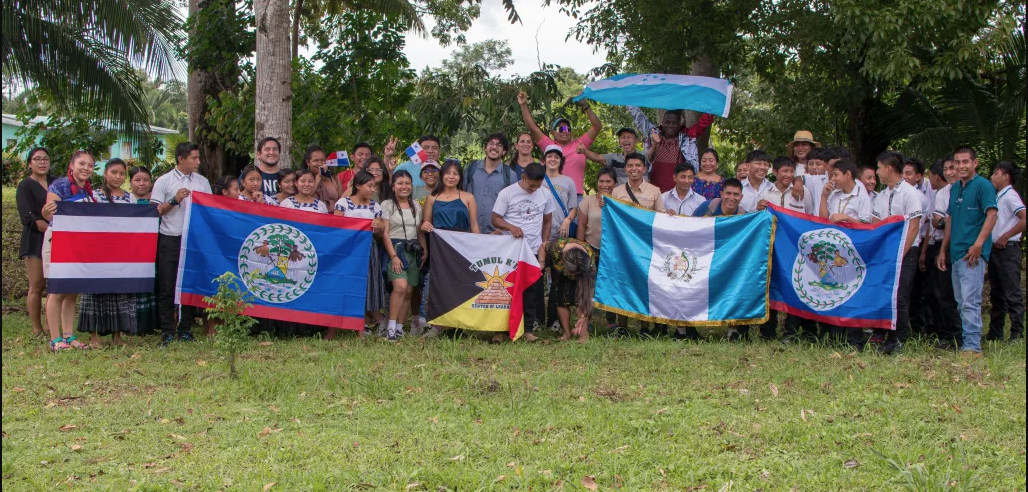 Un grupo de personas en un ambiente natural, con arbolead detras posando para la foto con banderas de diferentes paise.