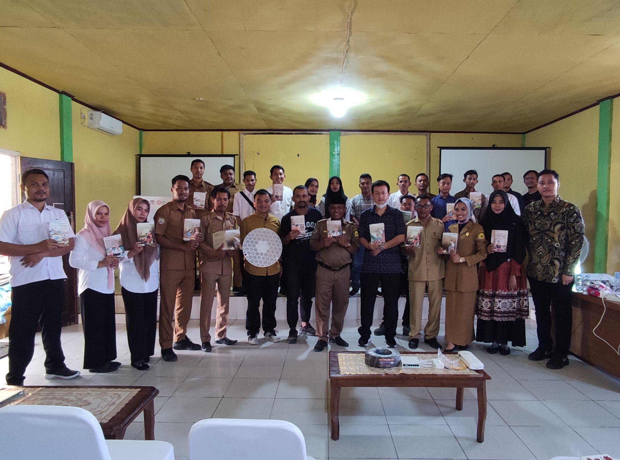 A group of 24 people stands in a room with yellow walls, holding books and a decorative plate. They are smiling, suggesting a joyful, celebratory event.