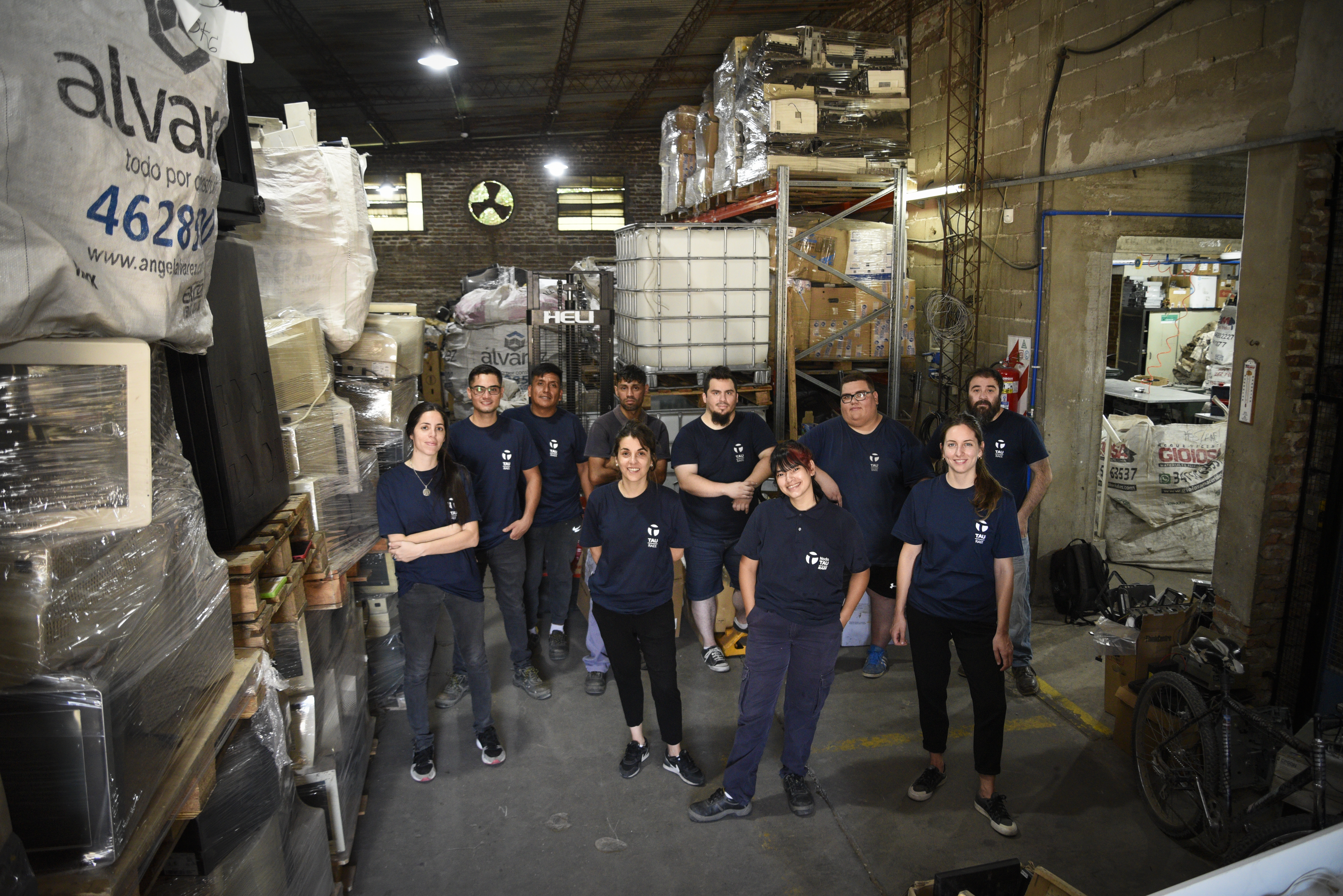 Grupo de jóvenes todos con la misma vestimenta, con el logo TAU, en un ambiente con equipos informáticos empaquetados