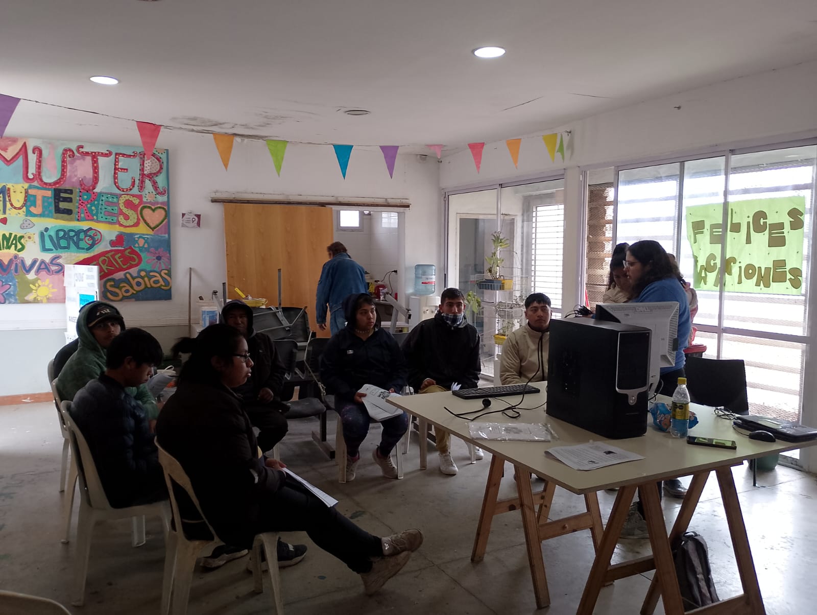 Grupo de personas alrededor de uan mesa con una computadora en una sala decorada con carteles coloridos y banderines