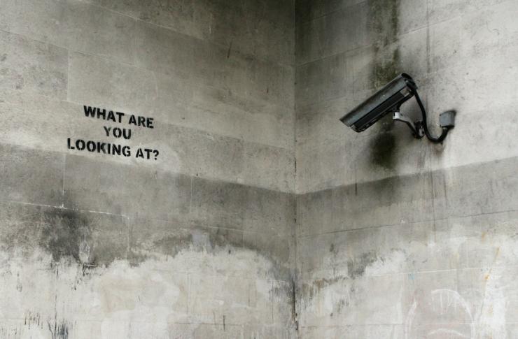 A camera points at a wall with graffiti that reads, ‘What are you looking at?’